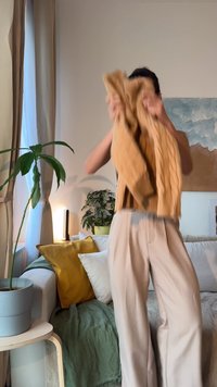 Beige kabelstrikket genser holdt opp, med et teksturert mønster, kombinert med off-white vide bukser, mot en koselig innendørs bakgrunn.