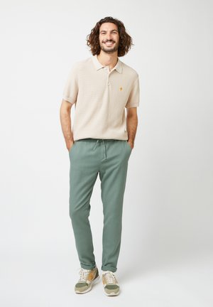 Beige textuur polo shirt met een lichte kraag, groene broek met trekkoord en grijze sneakers met oranje accenten. Model staat en lacht.