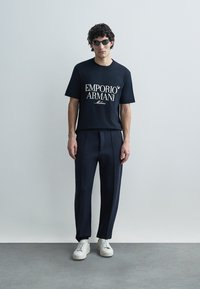 Svart t-shirt med vit "EMPORIO ARMANI Milano"-tryck, kombinerad med skräddarsydda mörka byxor och vita sneakers. Modellen bär mörka solglasögon.