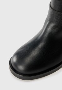 Botín de cuero negro hasta el tobillo con punta redonda, textura suave y suela resistente. Presenta una costura sutil cerca del eje.