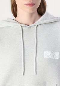 Lys grå hettegenser med en myk tekstur, med hvite snorer og trykt logo-tekst på forsiden i vertikal orientering.