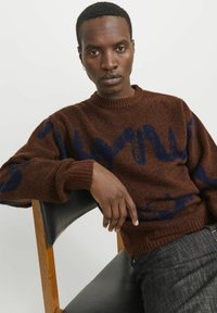 Brauner Strickpullover mit blauen abstrakten Mustern, runder Ausschnitt, gerippte Manschetten und Saum. Trägt dunkle Jeans und sitzt auf einem Stuhl.