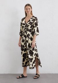Black and beige floral dress with a V-neck, knotted front, three-quarter sleeves, and side slits, paired with flat black sandals.