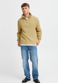 Bež kudumiga pullover, millel on pooltõmblukuga krae ja ribakujuline tekstuur, kantud valge särgi üle, kergete siniste teksadega ja mustade kingadega.