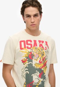 Camiseta de algodón beige que presenta un gráfico colorido de un tigre con olas y llamas, texto en negrita "OSAKA" en rojo y mangas cortas.