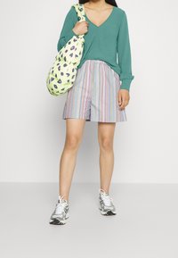 Žena oblečená v tmavě tyrkysovém topu s dlouhým rukávem, multibarevných pruhovaných šortkách, bílých teniskách, nese světle zelenou tašku přes rameno s leopardím vzorem.