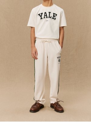 T-shirt crème avec l'inscription "YALE" en lettres noires audacieuses, associé à un jogging crème avec des bandes latérales vertes et un logo. Chaussures marron à lacets.