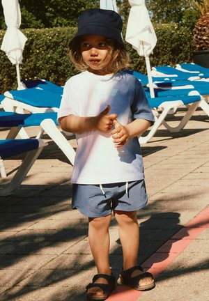Junge Kind trägt einen marineblauen Anglerhut, ein weiß-blaues Hemd und blaue Shorts und steht auf einer Terrasse am Pool in der Nähe von blauen Liegestühlen.