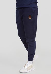 Námořnicky modré tepláky s žebrovanými manžetami a bočními kapsami. Na levém stehně je vyšívaný emblém. Měkká, texturovaná tkanina.