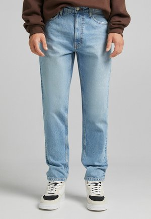 Person iført lyseblå jeans med lige ben, hvide og sorte sneakers og en brun langærmet top, stående mod en ensfarvet baggrund.