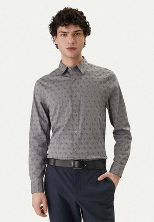 Giovane uomo che indossa una camicia grigia con motivo a bottoni infilata in pantaloni blu navy con cintura nera, in piedi con una mano in tasca.