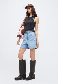 Γυναίκα με μαύρο τοπ χωρίς μανίκια, γαλάζια τζιν σορτς, μαύρες ψηλές μπότες, καφέ καπέλο, κρατώντας καφέ και λευκή τσάντα ώμου με σχέδιο.