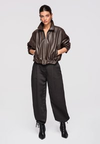 Femme portant une veste ceinturée en similicuir marron foncé, un pantalon noir ample et des bottines à talons noires, les mains dans les poches de la veste.