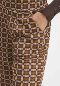 Un pantalon à carreaux marron présente un mélange de motifs marron, jaune et blanc, avec des poches latérales et un tissu texturé. Détail avec une taille côtelée.