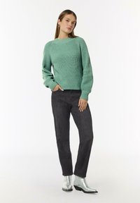 Mintgrüner gestrickter Pullover mit gerippter Textur, kombiniert mit dunkelgrauen Slim-Fit-Jeans. Das Modell trägt silberne Ankle-Boots und schlichte Ohrringe.