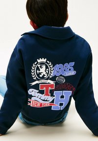 Bambino che indossa un felpa blu navy con grandi grafiche colorate "Tommy," "classe del 1985," stemma del leone e "New York" sul retro.