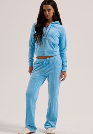 Vrouw die een lichtblauwe velours hoodie met rits en bijpassende trainingsbroek draagt, staand met één hand ontspannen langs haar zijde.