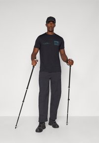 Camiseta negra de manga corta con gráfico de montaña azul, combinada con pantalones de senderismo grises, sosteniendo bastones de trekking, usando una gorra negra y zapatos.