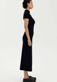 Czarna, dopasowana sukienka midi z krótkimi rękawami i wysokim kołnierzem. Gładki, elastyczny materiał o subtelnym połysku. Noszona z czarnymi klapkami.