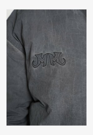 Sivá bunda s textúrovaným povrchom, s vyšívaným logom v tmavšej sivej na ľavej strane. Materiál pôsobí odolne a pevne.