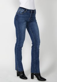 Femme portant un jean denim bleu avec des ourlets légèrement effilochés et des bottines noires à talons, debout sur un fond blanc.