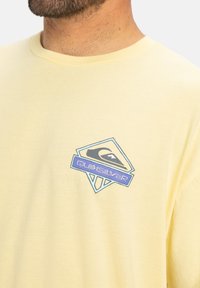 Krátký rukáv žlutého trička vyrobeného z bavlny, s vícebarevným logem Quiksilver na levé hrudi. Hladká textura, uvolněný střih.