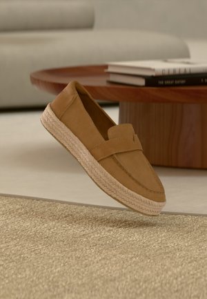 Chaussure à enfiler couleur tabac avec une semelle en jute tressée, présentée flottant au-dessus d'un tapis beige tissé dans un salon.