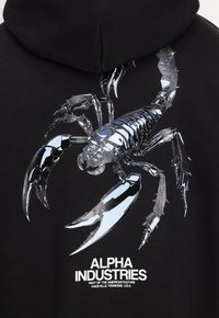 Sweat à capuche noir avec un motif de scorpion métallique argenté dans le dos, accompagné du texte "ALPHA INDUSTRIES" en blanc en dessous.