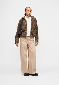 Νεαρή γυναίκα με πλεξούδες, που φοράει καφέ μπουφάν με χνουδωτή υφή, λευκό πουκάμισο, μπεζ παντελόνι με φαρδιά μπατζάκια, μαύρες μπότες και μαύρη τσάντα χιαστί.