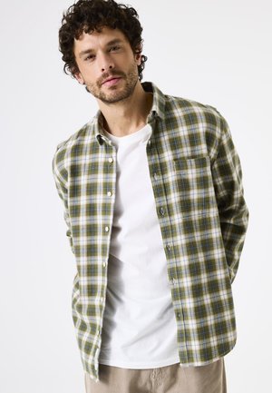 Chemise en flanelle à carreaux verts et beiges avec un col boutonné. Portée par-dessus un T-shirt blanc, elle dispose d'une poche poitrine et d'une texture douce.