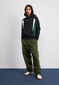 Mustapaita, jossa on vihreitä ja valkoisia yksityiskohtia, logolla varustettuna. Oliivinvihreät ripeät housut ja valkoiset kengät, joissa on vaahtokumipohja.