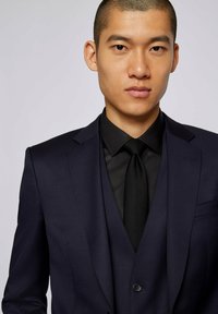 Granatowa marynarka z wyciętymi klapami, czarna koszula i krawat. Gładki materiał o dopasowanym kroju. Detal z guzikami na klapie marynarki.