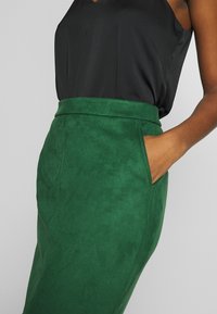 Grön mockapennkjol med hög midja, sidofickor och en slät yta. Har en strukturerad design och en subtil lyster.