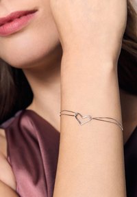 Srebrna bransoletka z charmsami w kształcie serca, delikatne łańcuszki, gładka tekstura, noszona na nadgarstku o jasnej karnacji, na tle ciemnego materiału.