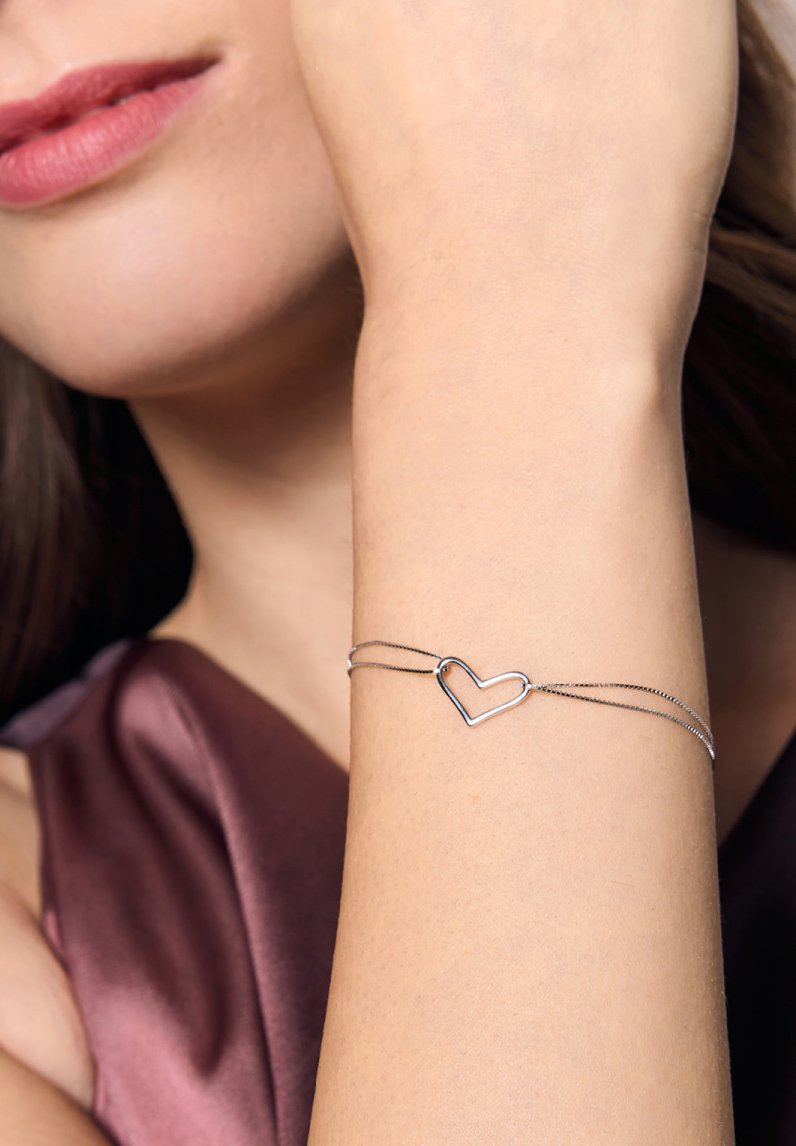 Srebrna bransoletka z charmsami w kształcie serca, delikatne łańcuszki, gładka tekstura, noszona na nadgarstku o jasnej karnacji, na tle ciemnego materiału.