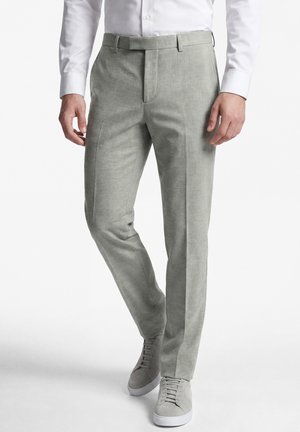 Man draagt lichtgrijze nette broek gecombineerd met bijpassende grijze sneakers en een wit shirt met lange mouwen, staand tegen een witte achtergrond.
