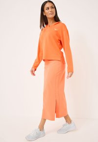 Oransje cropped hettegenser med lange ermer, parret med en matchende midi-skjørt med en sidesliss. Brukt med lysegrå joggesko.