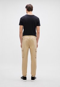 Beige cargobroeken met elastische boorden en zijzakken, gecombineerd met een zwart shirt met korte mouwen, gezien van achteren.