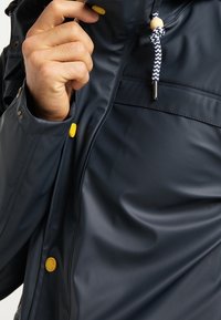 Veste imperméable bleu marine avec une texture lisse, des accents de boutons jaunes, et un col à cordon de serrage orné d'un cordon tressé.