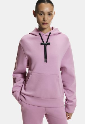 Žena v pastelovo ružovej mikine s kapucňou a k tomu ladiacich nohaviciach s čiernymi šnúrkami, stojaca s jednou rukou v vrecku na bielom pozadí.