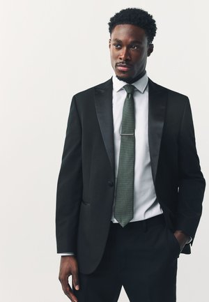 Mann in schwarzem Smoking mit Satinrevers, weißem Hemd und gemustertem grünen Krawatte, steht mit einer Hand in der Tasche und schaut seitlich vor einfarbigem Hintergrund.