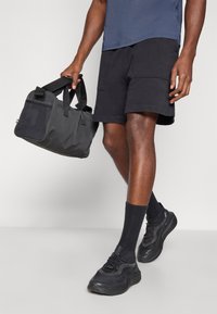 Saco duffel preto com alças duplas e painel lateral em malha, combinado com calções de atletismo pretos, meias pretas e sapatilhas pretas.