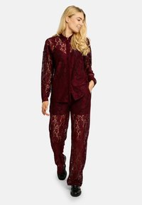 Vinovočervený dvojdielny súbor z čipky, ktorý obsahuje košeľu na gombíky s dlhými rukávmi a nohavice s širokými nohavicami, oba s kvetinovými vzormi a priehľadnou textúrou.