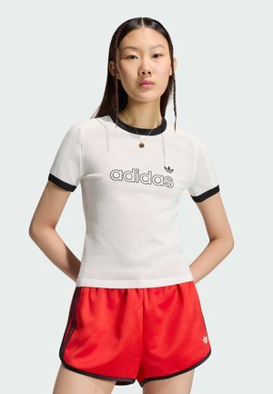Mladá žena nosí bílý síťovaný Adidas tričko s černým lemováním a červené Adidas šortky, stojí s rukama za zády.