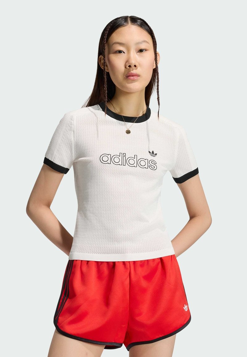 Jeune femme portant un t-shirt en mesh blanc Adidas avec bordure noire et un short rouge Adidas, debout les mains derrière le dos.