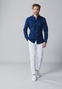 Marineblauwe button-up shirt, witte slim-fit broek en witte sneakers, staand tegen een neutrale achtergrond. Glad stof met een klassieke kraag.