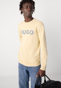 Lys gul hettegenser med "HUGO" i grå bokstaver på forsiden, kombinert med blå denimjeans. Stoffet ser mykt og jevnt ut.