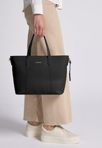 Schwarze Leder-Shopper-Tasche mit strukturierter Form, doppelten Griffen und einem goldenen Logo-Akzent. Getragen von einer Person in beigen Hosen und weißen Sneakers.