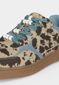 Chaussure de sport avec imprimé vache marron et beige, lacets bleus enfilés de perles blanches, détails en daim bleu et semelle marron clair.