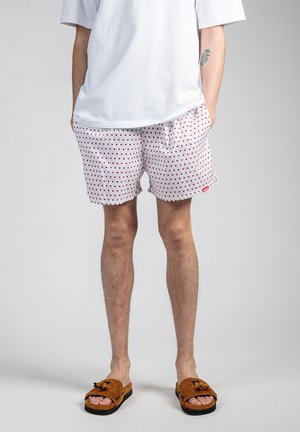 Hvide badebukser med røde hjerte mønstre, elastisk talje, to sidelommer, parret med brune teksturerede sandaler. Ensfarvet hvid t-shirt.